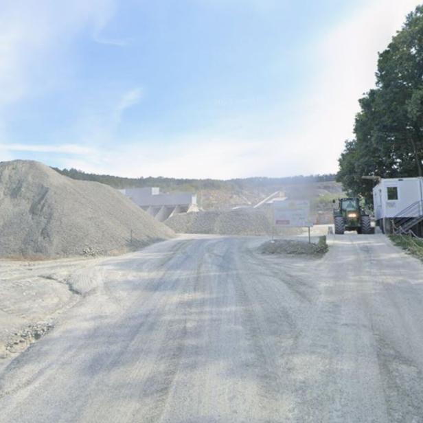 Große Schotterhaufen, ein Traktor und Container stehen auf einem staubigen Gelände neben Bäumen unter blauem Himmel.
