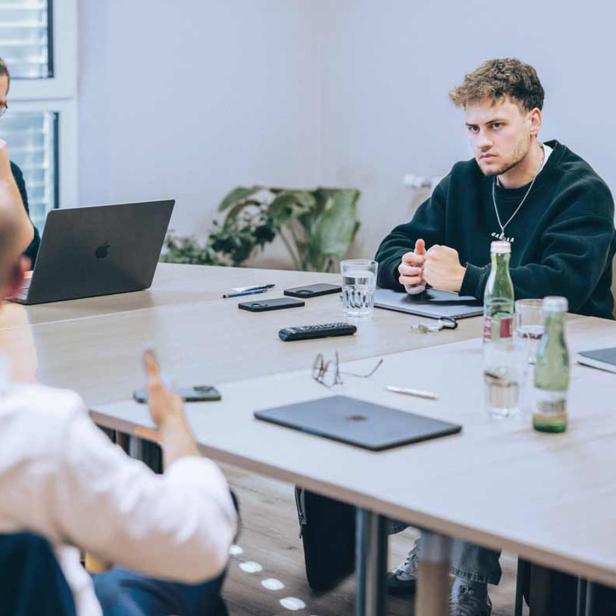 Vier Männer sitzen an einem Tisch und führen eine konzentrierte Besprechung mit Laptops, Getränken und Notizbüchern.