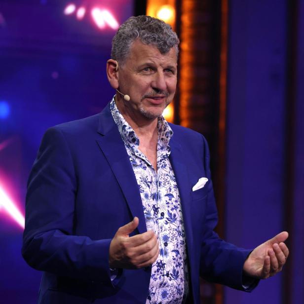 Ein Mann mit Mikrofon im blauen Anzug gestikuliert vor farbigem Hintergrund.