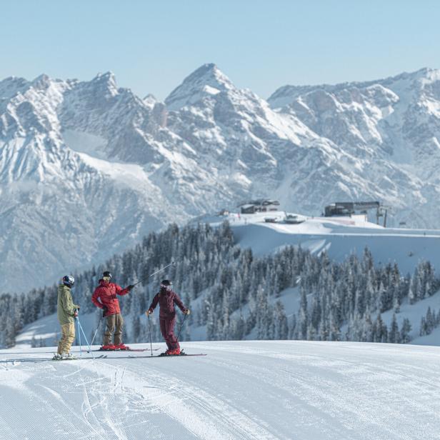 Drei Skifahrer stehen auf einer verschneiten Piste vor einer Kulisse aus schneebedeckten Bergen.