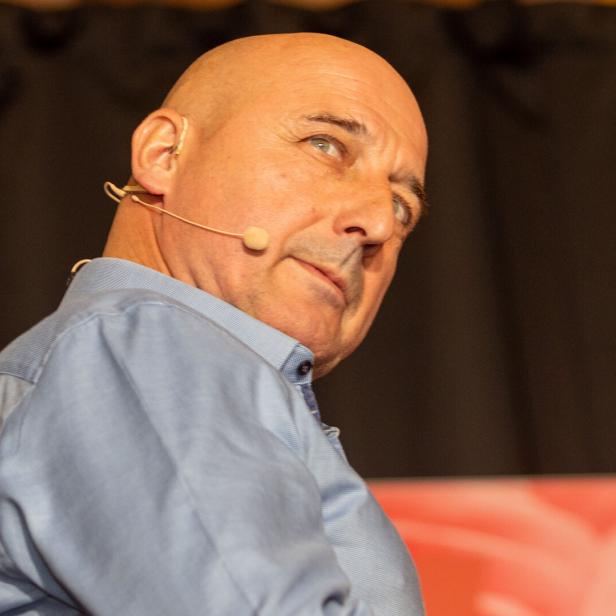 Ein Mann mit Glatze und blauem Hemd trägt ein Headset-Mikrofon und blickt nach oben.