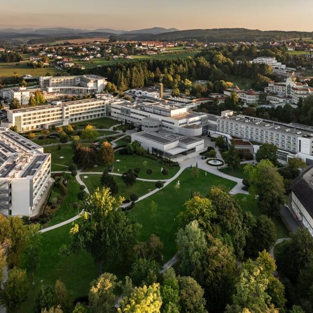 Mehrere moderne Gebäude mit weißen Fassaden liegen inmitten einer weitläufigen, grünen Parkanlage mit Wegen und Bäumen.