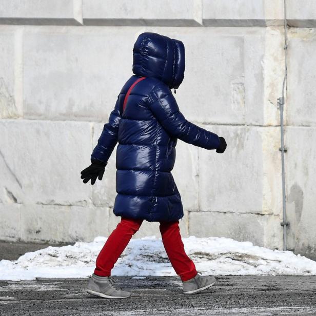 Person mit langer, dicker, blauer Daunenjacke, roter Hose und grauen Schuhen geht an einer schneebedeckten Straße entlang.