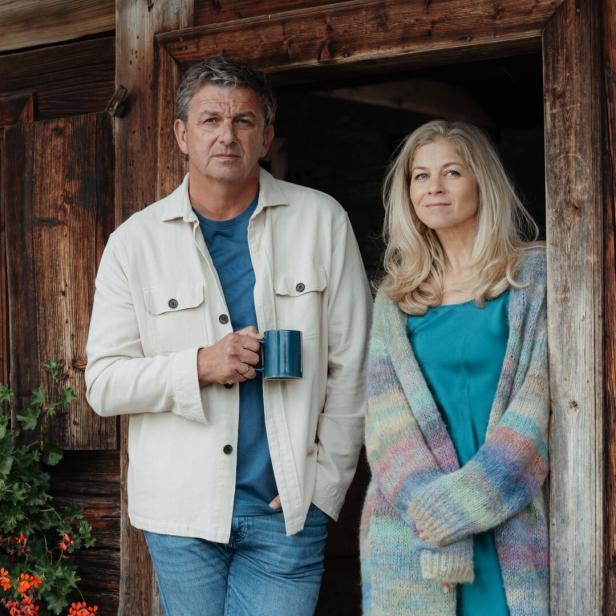 Ein Mann mit Tasse und eine Frau stehen entspannt an einer Holzhütte neben roten Blumen.