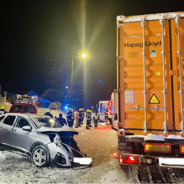 Ein silbernes Auto ist frontal mit einem orangefarbenen Lkw kollidiert, Feuerwehrleute und Einsatzfahrzeuge sind vor Ort.
