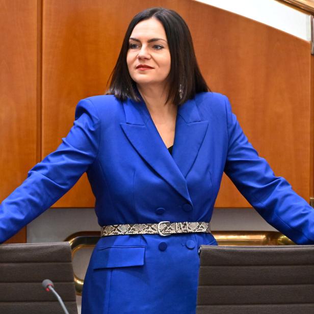 Frau mit schulterlangem dunklem Haar trägt einen blauen Blazer und steht mit ausgebreiteten Armen an zwei Stühlen.