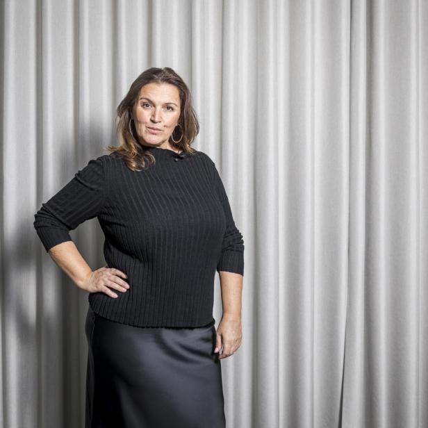 Frau mit langen braunen Haaren trägt schwarzen Pullover und Rock, steht vor einem hellgrauen Vorhang.