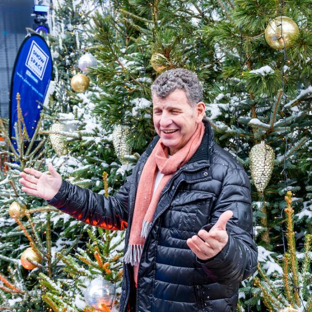 Ein Mann mit schwarzer Jacke und rotem Schal steht lachend zwischen geschmückten, verschneiten Tannenbäumen.