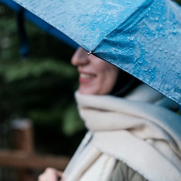 Person mit Schal und blauem Regenschirm im Regen, unscharfer Hintergrund mit Grün.