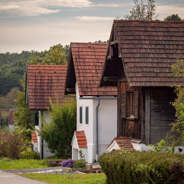 Mehrere kleine Häuser mit roten Ziegeldächern stehen nebeneinander an einer ruhigen Straße, umgeben von viel Grün.