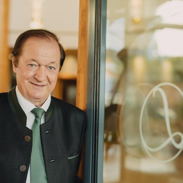 Ein älterer Mann im Trachtenanzug mit grünem Schlips steht lächelnd neben einer Glastür mit verziertem Logo.