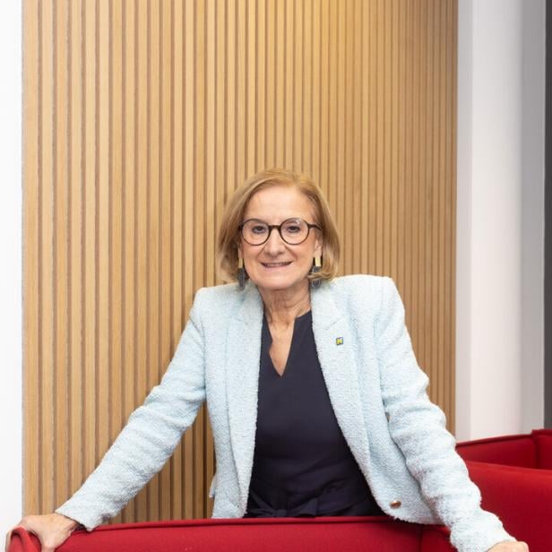 Eine Frau mit Brille und hellblauem Blazer steht lächelnd hinter einer roten Sitzgelegenheit vor einer Holzvertäfelung.