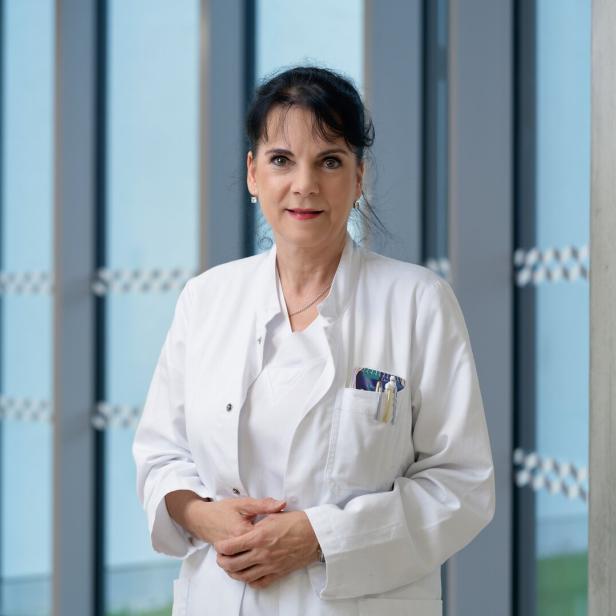 Eine Frau im weißen Arztkittel steht mit Stiften in der Brusttasche vor großen Fenstern und blickt freundlich nach vorn.