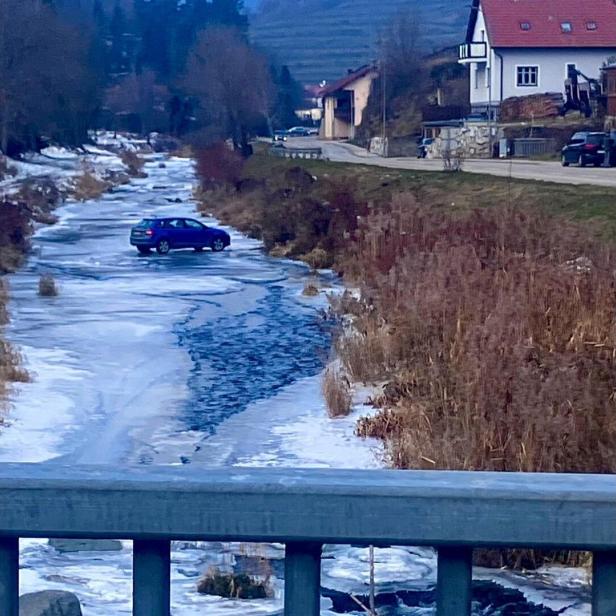 Ein blaues Auto steht mitten auf einem zugefrorenen Fluss, umgeben von winterlicher Landschaft und Häusern am Ufer.