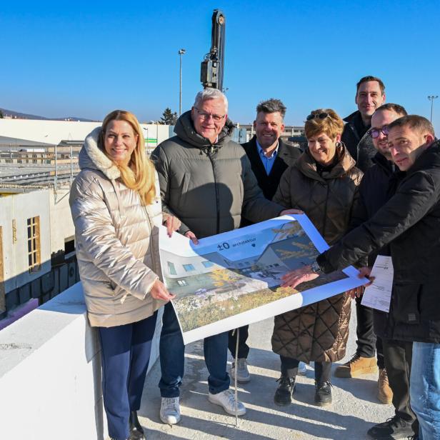Sieben Personen stehen auf einer Baustelle und betrachten gemeinsam einen großen Architekturplan.