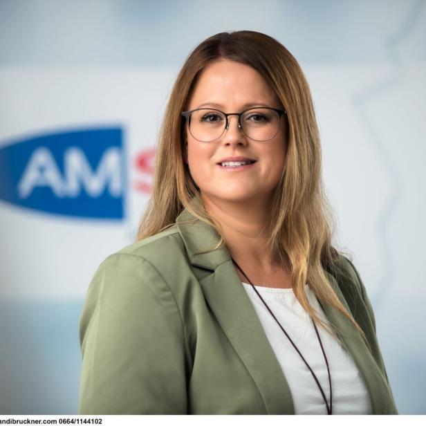 Frau mit langen blonden Haaren, Brille und grünem Blazer steht vor einem unscharfen Hintergrund mit blauem Logo.