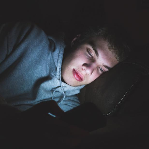 Ein junger Mann liegt im Dunkeln und blickt auf sein leuchtendes Handy.