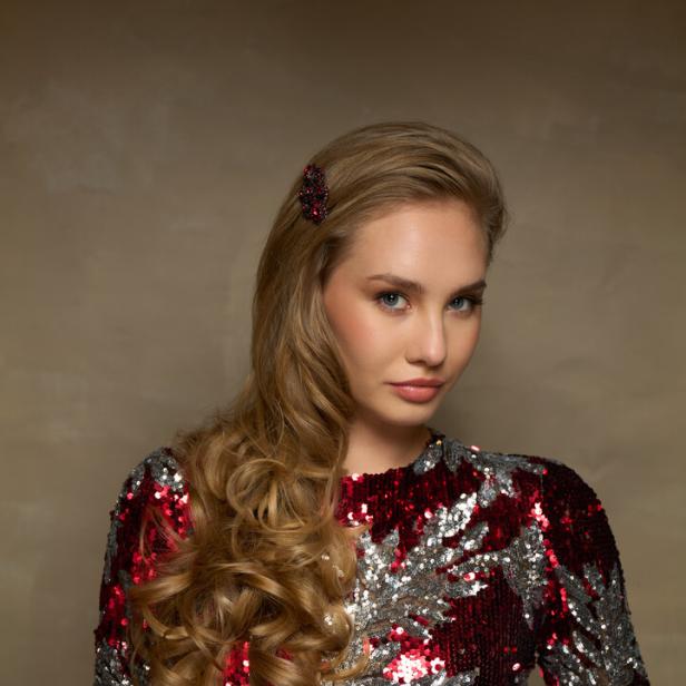 Eine Frau mit blonden Locken trägt ein glitzerndes, rot-silbernes Kleid und eine Haarspange.