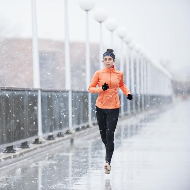 Eine Frau joggt bei Schneefall auf einer Brücke und trägt eine orange Jacke sowie schwarze Leggings.
