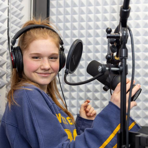 Eine junge Frau mit Kopfhörern lächelt in einem Tonstudio vor einem Mikrofon.