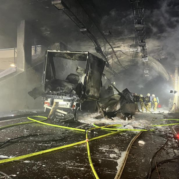 In einem Tunnel steht ein ausgebrannter LKW, umgeben von Löschschaum und Feuerwehrleuten.