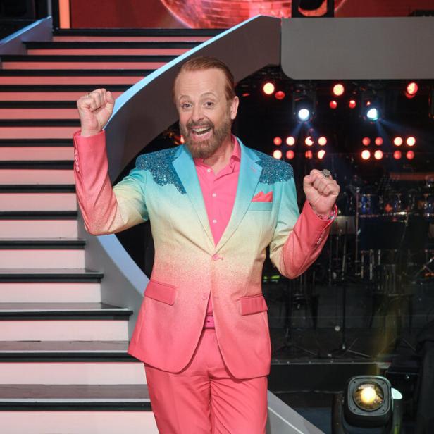 Ein Mann mit Bart trägt einen auffälligen Anzug in Pink und Türkis und posiert fröhlich vor einer beleuchteten Showtreppe.