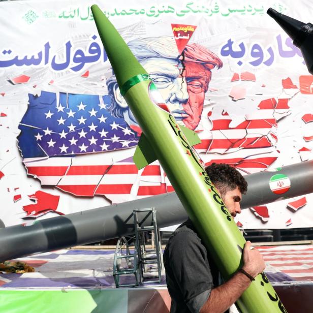 Ein Mann trägt eine grüne Rakete vor einem Banner mit zerrissenen Flaggen der USA und Israels.