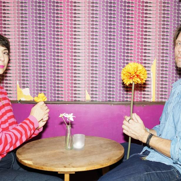 Zwei Personen (Ruth Brauer-Kvam und Kyrre Kvam) sitzen sich an einem kleinen runden Tisch gegenüber und halten jeweils eine gelbe Blume in der Hand.