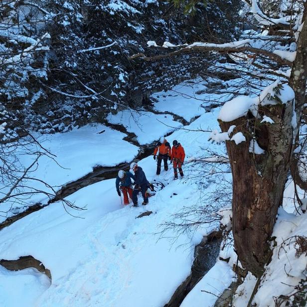 Eine Gruppe von Bergrettern überquert einen schneebedeckten Bach in einer bewaldeten Umgebung.