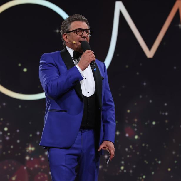 Ein Mann im blauen Anzug spricht in ein Mikrofon vor einem dunklen Hintergrund mit Lichtern.