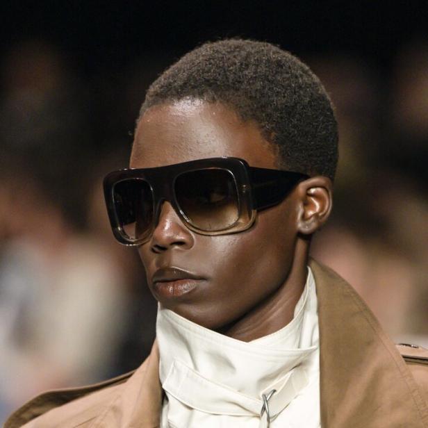 Person mit großer, dunkler Sonnenbrille, kurzem Haar, beigem Mantel und weißem, hochgeschlossenem Oberteil bei der Pariser Fashion Week