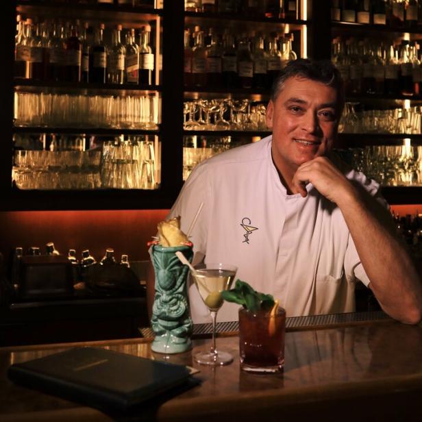 Barchef Heinz Kaiser lächelt hinter seiner Bar mit drei seiner Mocktails und vollen Spirituosenregalen.