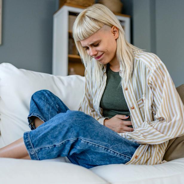Eine junge Frau sitzt auf einem Sofa und hält sich mit schmerzverzerrtem Gesicht den Bauch.