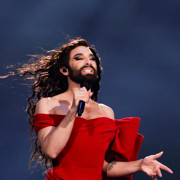 Conchita Wurst singt in einem roten Kleid mit Bart und wallendem Haar.