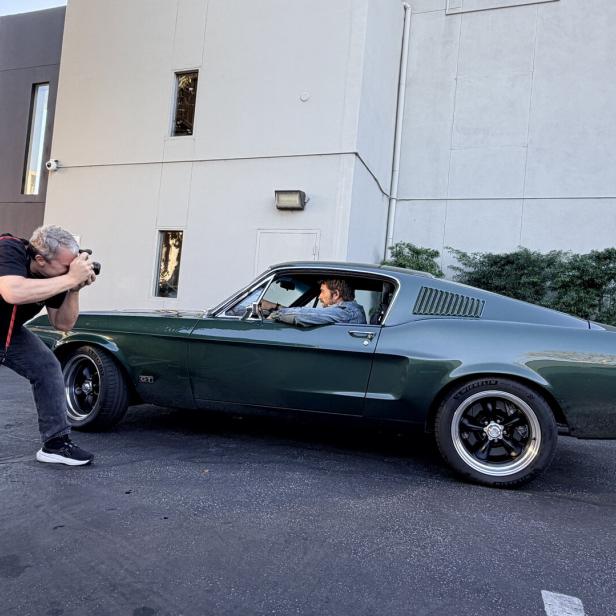 Ein Fotograf macht ein Bild von einer Person, die in einem dunkelgrünen Oldtimer vor einem Gebäude sitzt.