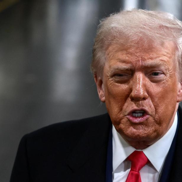 Donald Trump spricht mit roter Krawatte und dunklem Anzug vor grauem Hintergrund.