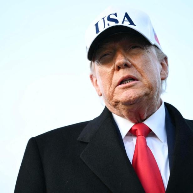 Donald Trump mit "USA"-Kappe, dunklem Anzug und roter Krawatte vor hellem Hintergrund.