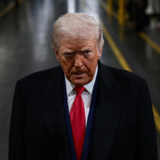 Donald Trump blickt ernst in die Kamera, gekleidet in einen dunklen Mantel und eine rote Krawatte.