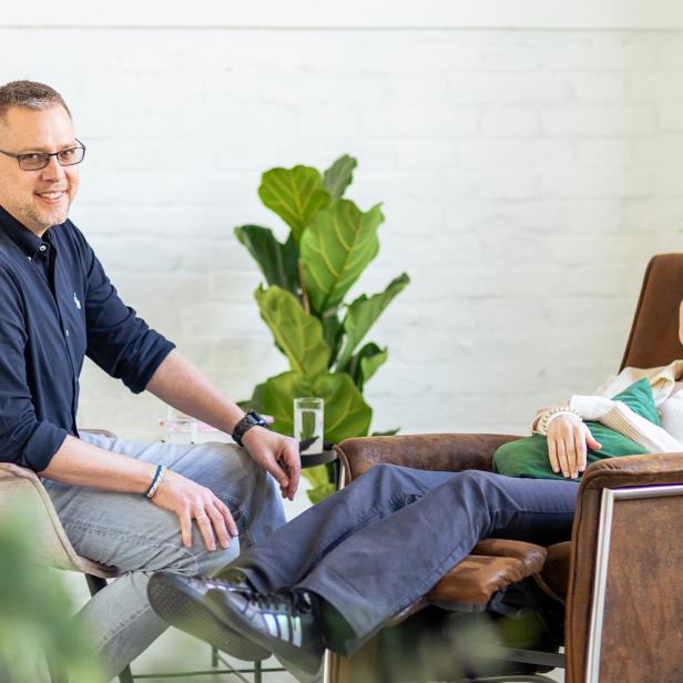 Ein Mann sitzt neben einer entspannt liegenden Frau in einem hellen Praxisraum mit Pflanzen.