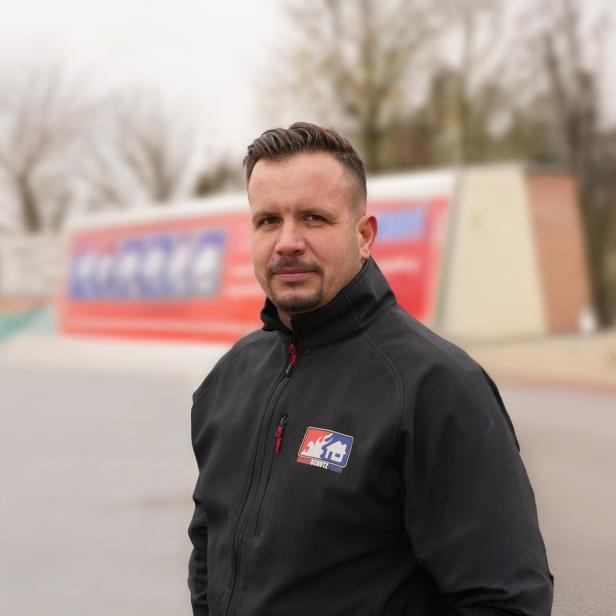 Mann in schwarzer Jacke mit Brandschutz-Logo steht im Freien vor unscharfem Hintergrund.