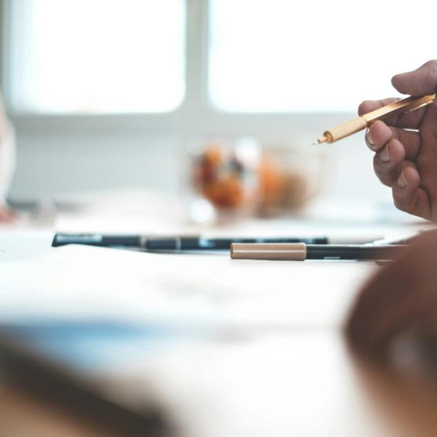 Zwei Personen sitzen an einem Tisch mit Stiften und Papier, eine Hand hält einen Kugelschreiber.
