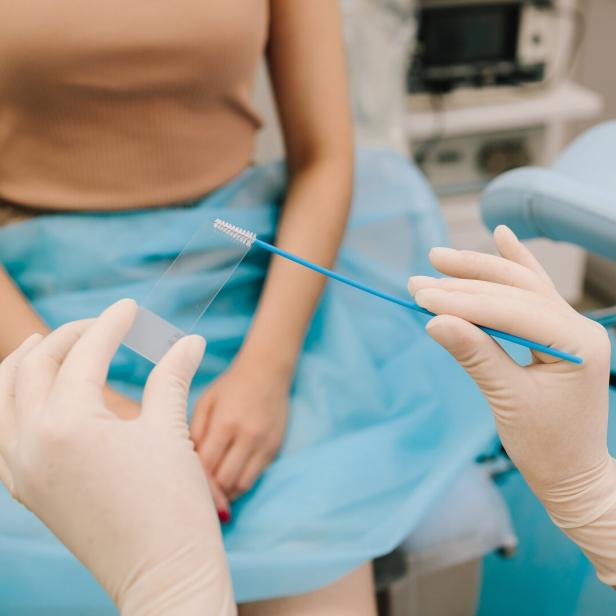 Eine Person mit Handschuhen hält einen Abstrich und einen Objektträger vor einer sitzenden Frau in medizinischer Umgebung.