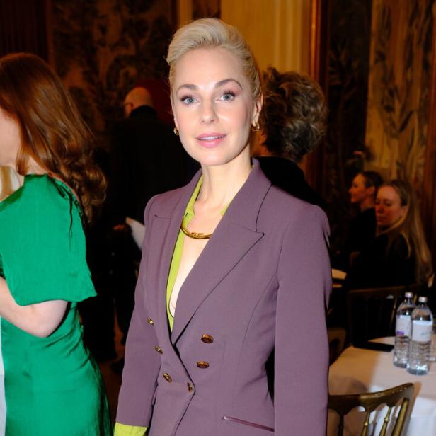 Eine Frau mit blondem Haar trägt einen lila Hosenanzug mit grünem Hemd und hält eine schwarze Handtasche.