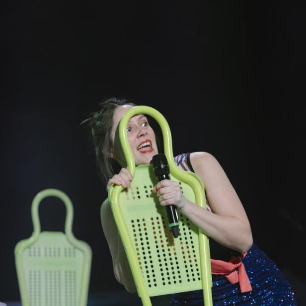 Frau in glitzerndem Kleid hält ein Mikrofon und einen grünen Stuhl, während sie auf einer Bühne steht.