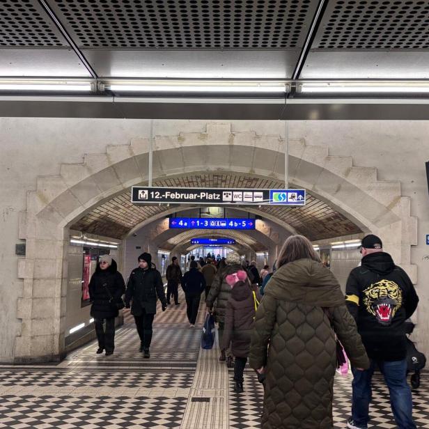 Menschen gehen durch einen Tunnel mit gemustertem Boden und Hinweisschildern zu Bahnsteigen und Ausgängen.