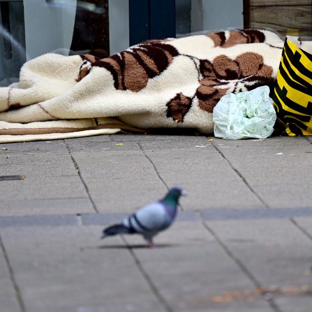 Eine Person liegt unter einer Decke auf dem Gehweg, daneben stehen eine Plastiktüte und eine Einkaufstasche, im Vordergrund Tauben.
