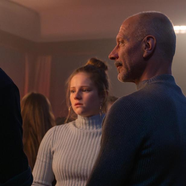 Drei Personen stehen in einem Raum, eine junge Frau in der Mitte mit besorgtem Blick.