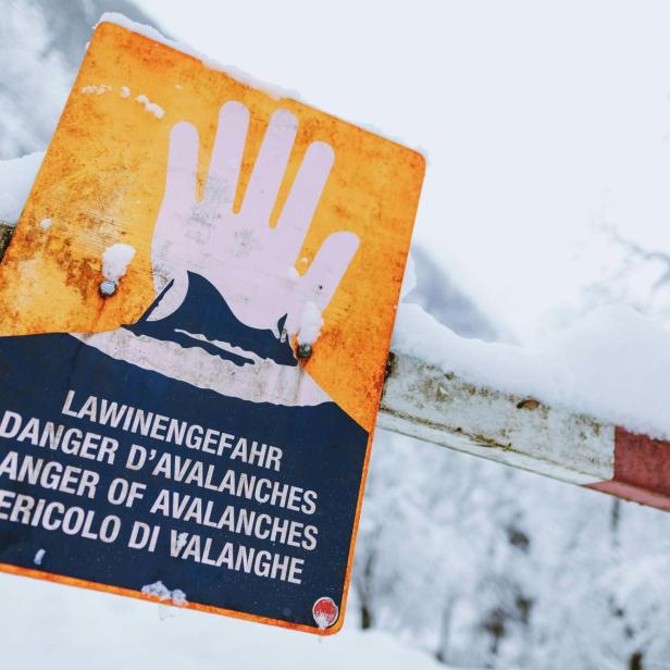 Warnschild mit erhobener Hand und Hinweis auf Lawinengefahr in mehreren Sprachen, umgeben von Schnee.