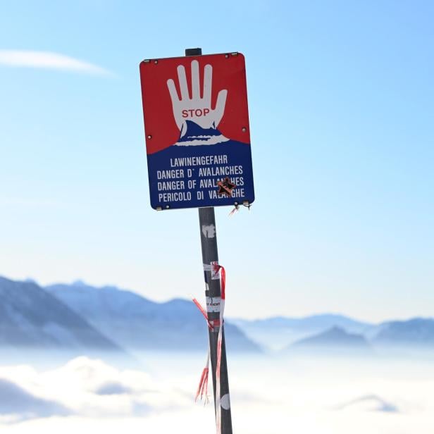 Warnschild mit roter Hand und Hinweis auf Lawinengefahr vor verschneiten Bergen bei klarem Himmel.