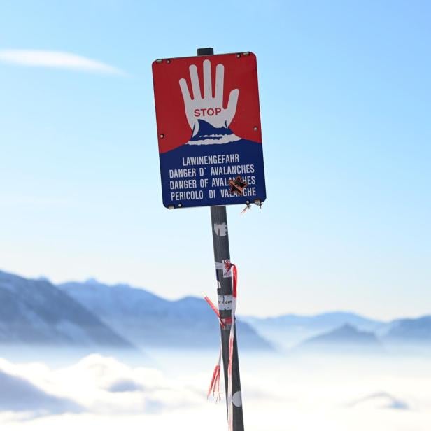 Warnschild mit roter Hand und Hinweis auf Lawinengefahr vor verschneiten Bergen bei klarem Himmel.